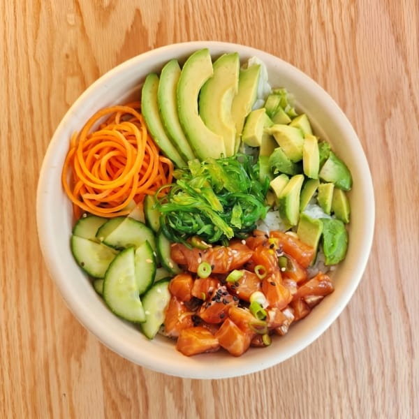 A colorful poke bowl with salmon, avocado, seaweed, carrots, and cucumber on a wooden table.