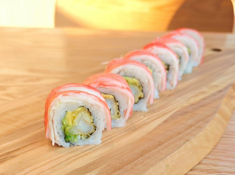 A row of crab and avocado sushi rolls on a light wooden cutting board.