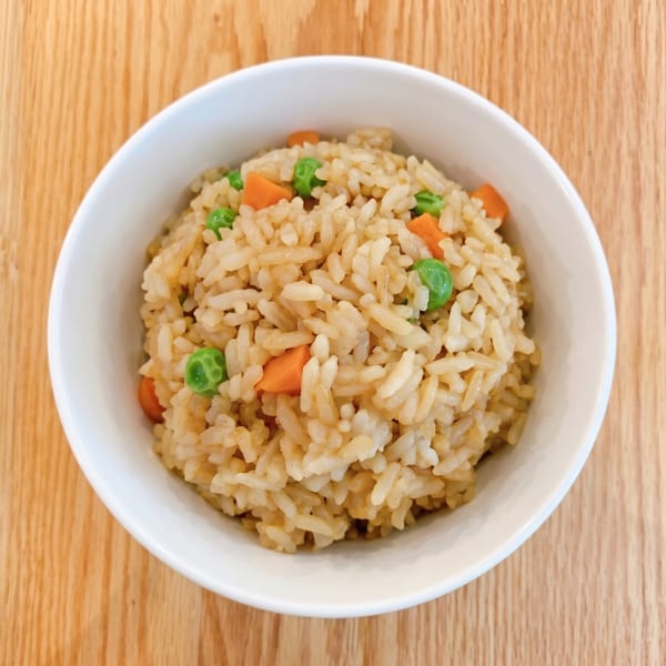 A white bowl filled with fried rice, green peas, and diced carrots on a wooden table.