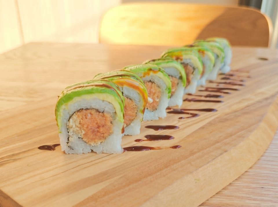 A row of sushi rolls with avocado and spicy salmon filling on a wooden board with sauce.