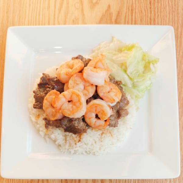A white rectangular plate holds rice, beef, shrimp, and shredded cabbage on a wooden table.
