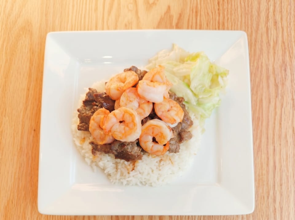 A white rectangular plate holds rice, beef, shrimp, and shredded cabbage on a wooden table.
