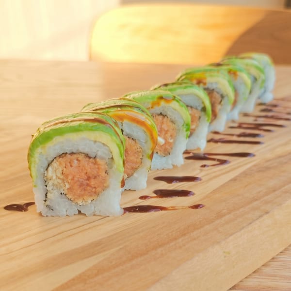 A row of sushi rolls with avocado and spicy salmon, drizzled with sauce on a wooden board.