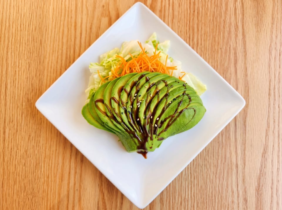 Avocado salad with shredded lettuce, carrots, balsamic glaze, and sesame seeds on a white plate.