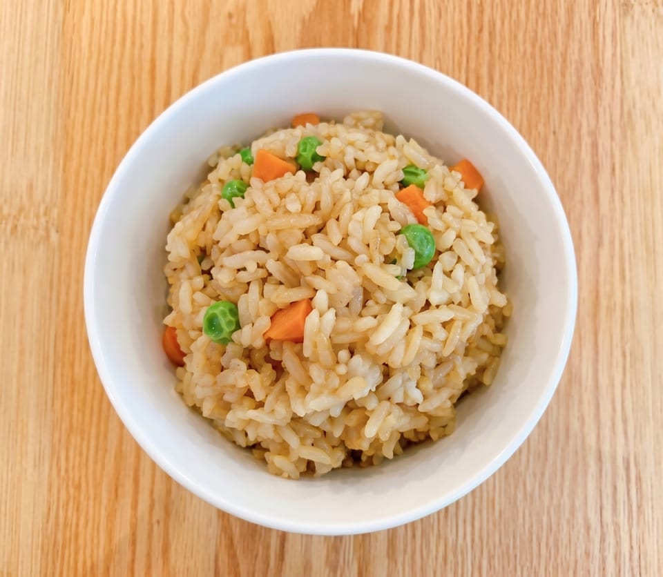 A white bowl filled with fried rice, green peas, and diced carrots on a wooden table.